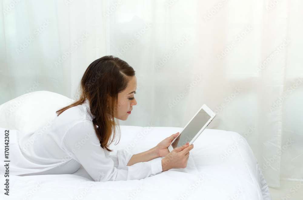 happy young asian woman in white pajamas playing mobile tablet on bed in the morning at home, soft focus, lifestyle smart communication technology, online shopping and payment online concept happy young asian woman in white pajamas playing mobile tablet on bed in the morning at home, soft focus, lifestyle smart communication technology, online shopping and payment online concept