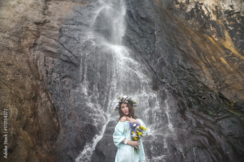 portrait of a girl with a wreath on her head and a bouquet of flowers in her hands against a waterfall