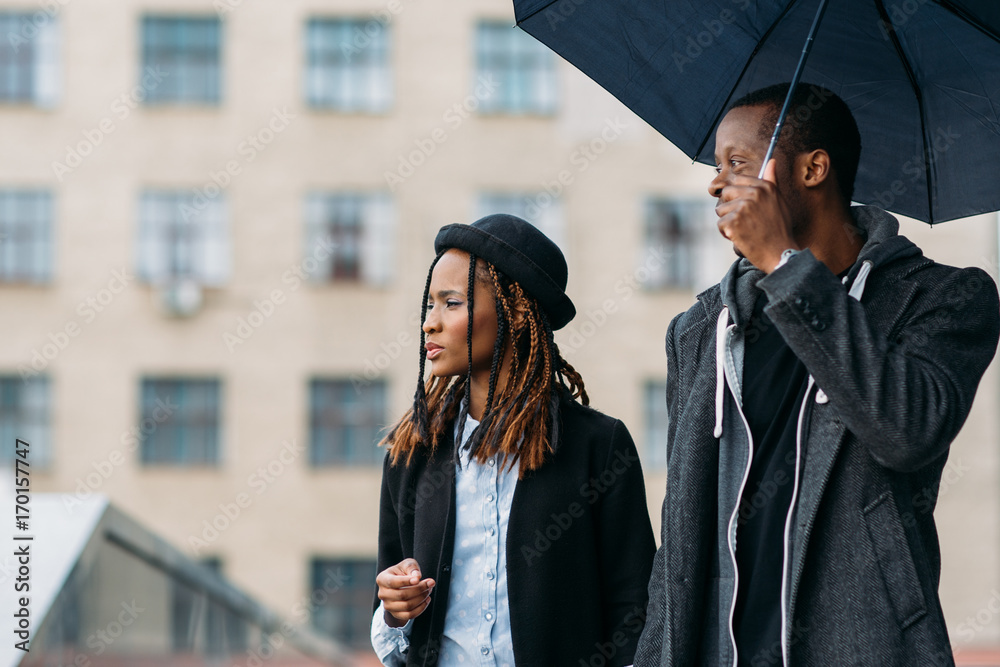 Thoughtful fashion pedestrians. Rainy mood. Young black couple with umbrella , happy African American, perspective concept