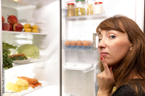 Confused young woman looking in fridge at kitchen