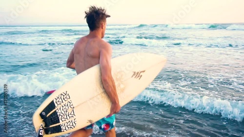 Young surfer holding board, running to go in the water at amazing golden light sunset. Happy man enjoying summer evening and seaside vacation activities. Shot taken by a handheld gimbal in 60FPS.