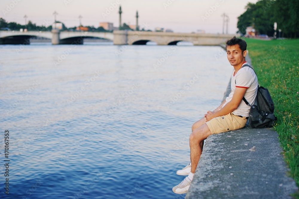 A young handsome guy sitting resting on the concrete Bank of embankment, lowering his feet over water, in summer on blurred background bridge and urban landscape.