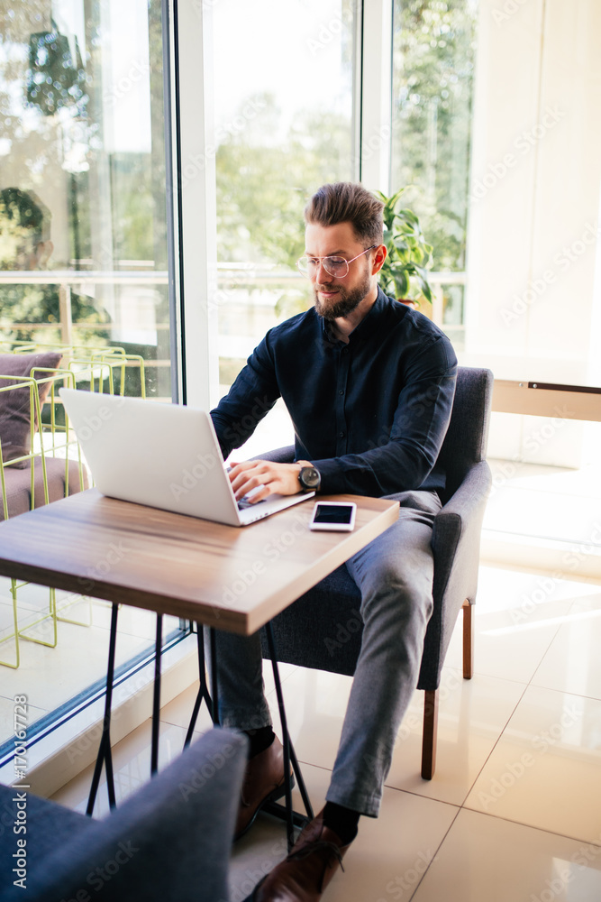 Successful young businessman typing on laptop at workplace big window on background