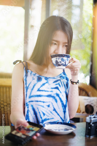 Portrait of young asian cute woman playing smart phone and testing a hot cup of coffee. in coffee cafe with vintage ceramic cup. selective focus. filtered image to vivid color. light effect added