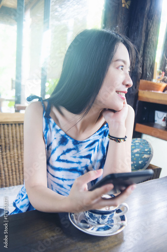 Portrait of young asian cute woman playing a smart phone and smiling. in coffee cafe with vintage ceramic cup. selective focus. filtered image to vivid color tone process. light effect added