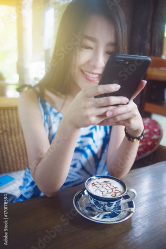Portrait of young asian cute woman playing a smart phone and smiling. in coffee cafe with vintage ceramic cup. selective focus. filtered image to vivid color tone process. light effect added