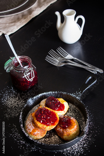 Still life with Four homemade fried Pancakes in small cast-iron frying pan. Raspberry jam in jar. Black wood background.