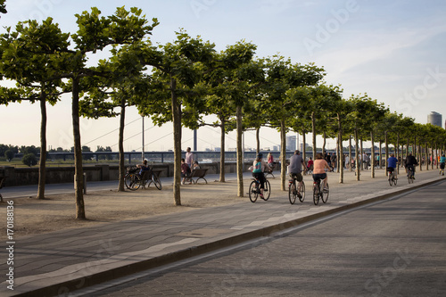 People ride bicycles by Rhine (Rhein) river. Tree line is also in the view. Image communicates lifestyle and culture of Dusseldorf.