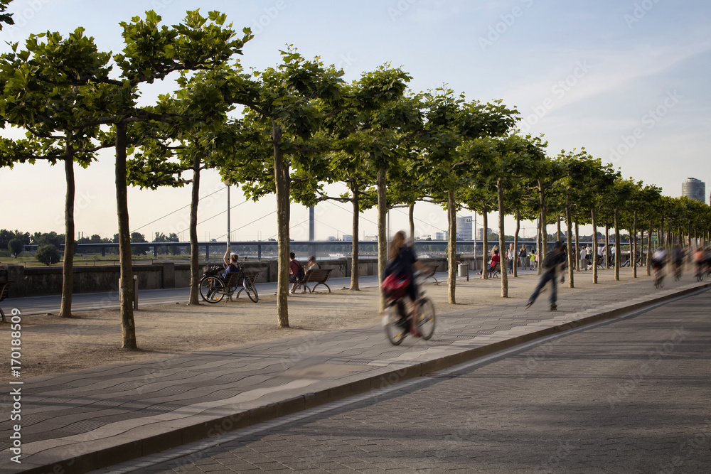 Woman rides bicycle and a boy rides skateboard in blurry motion by Rhine (Rhein) river. Tree line is also in the view. Image communicates lifestyle and culture of Dusseldorf.