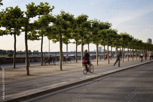 Woman rides bicycle and a boy rides skateboard in blurry motion by Rhine (Rhein) river. Tree line is also in the view. Image communicates lifestyle and culture of Dusseldorf.