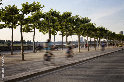 People ride bicycle in blurry motion by Rhine (Rhein) river. Tree line is also in the view. Image communicates lifestyle and culture of Dusseldorf.