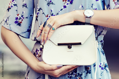 Close up photo of white leather bag in hands of fashionable woman pising in street. Model wearing silvery wrist watch, a lot of rings. Elegant outfit. Female fashion concept photo