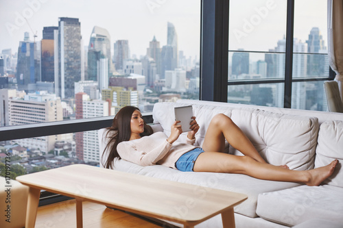 Asian young model lying and relax on the sofa with tablet in room with panoramic view to the modern city