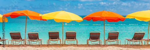 Fototapeta Naklejka Na Ścianę i Meble -  Beach chairs and umbrellas vacation tropical Holiday travel Banner. Caribbean beach on Sint Maarten with retro colorful parasols and loungers, turquoise blue ocean water. Panoramic crop.