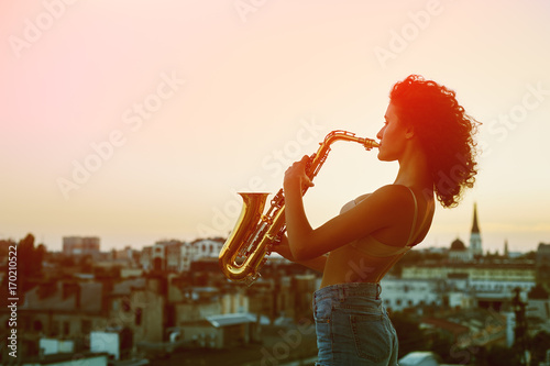 Canvas Print Young woman with saxophone on the roof of a high building