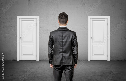 A businessman with his back turned stands between two identical closed white doors. photo