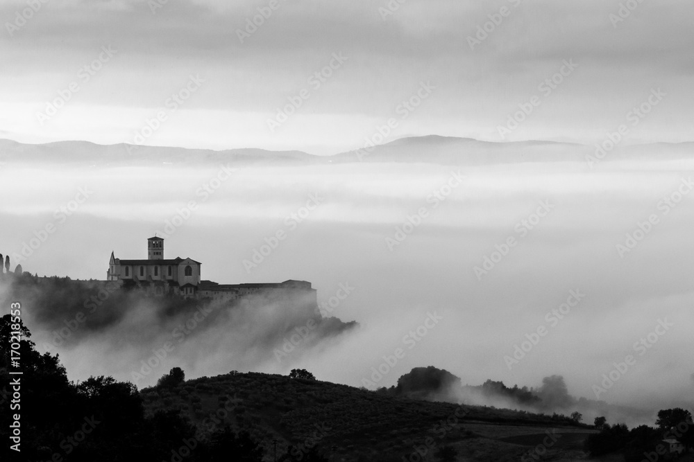 Beautiful monochrome view of St.Francis church in Assisi (Umbria, Italy), over a sea of fog at dawn, with hills and trees in the foreground
