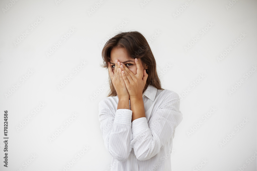 Mysterious enigmatic Hispanic female peeping out through her fingers. Picture of anxious young Latin woman employee wearing white blouse covering face with both hands, feeling terrified and scared Mysterious enigmatic Hispanic female peeping out through her fingers. Picture of anxious young Latin woman employee wearing white blouse covering face with both hands, feeling terrified and scared