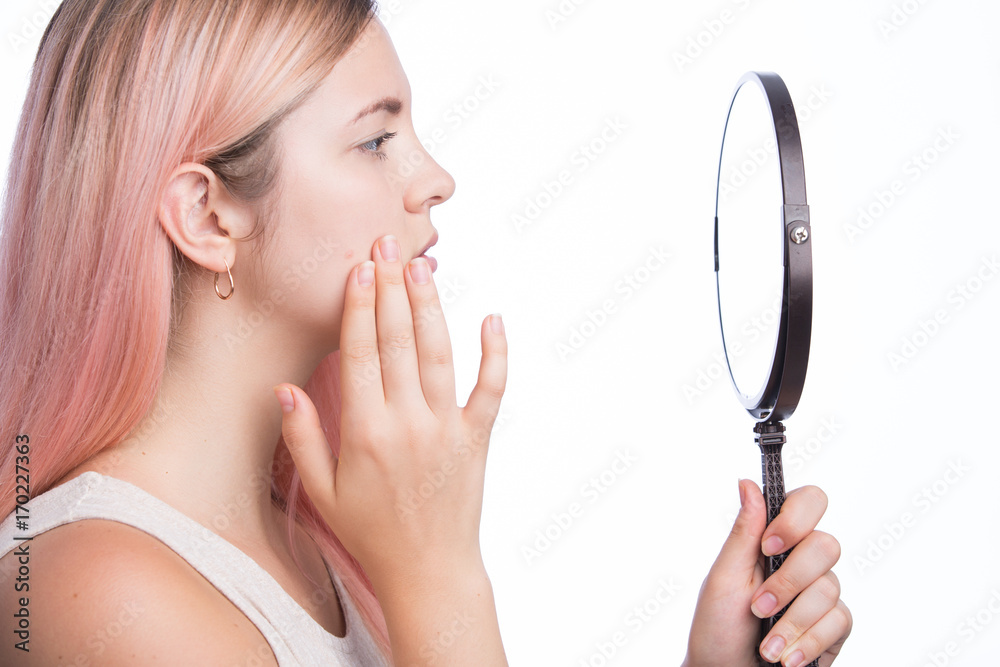 Unhappy young pretty girl looking at her pimple on her face in the mirror Unhappy young pretty girl looking at her pimple on her face in the mirror