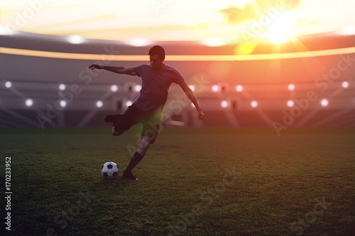 Soccer player is kicking a ball to the net in stadium at sunset.