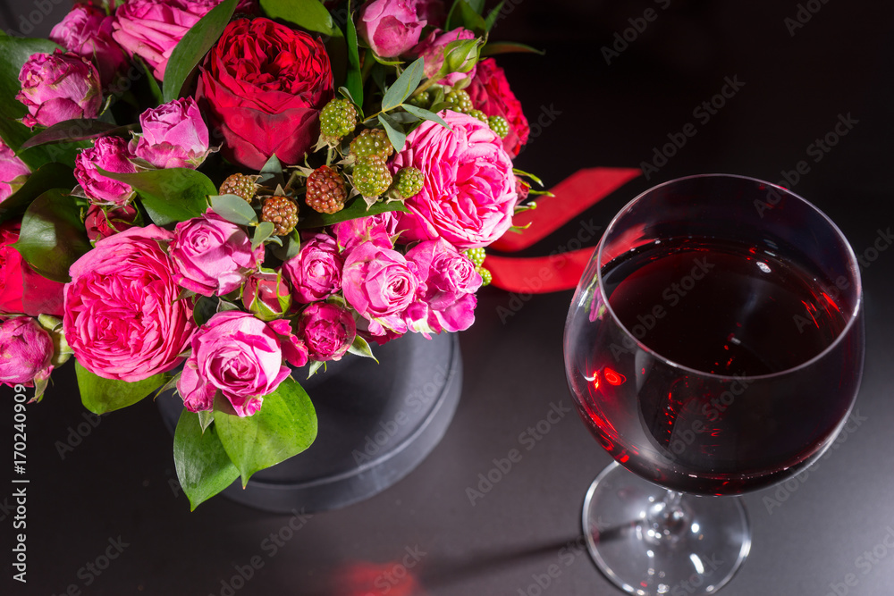 Stylish bouquet of pink and red roses and red ribbon in a circular black box near a glass with wine