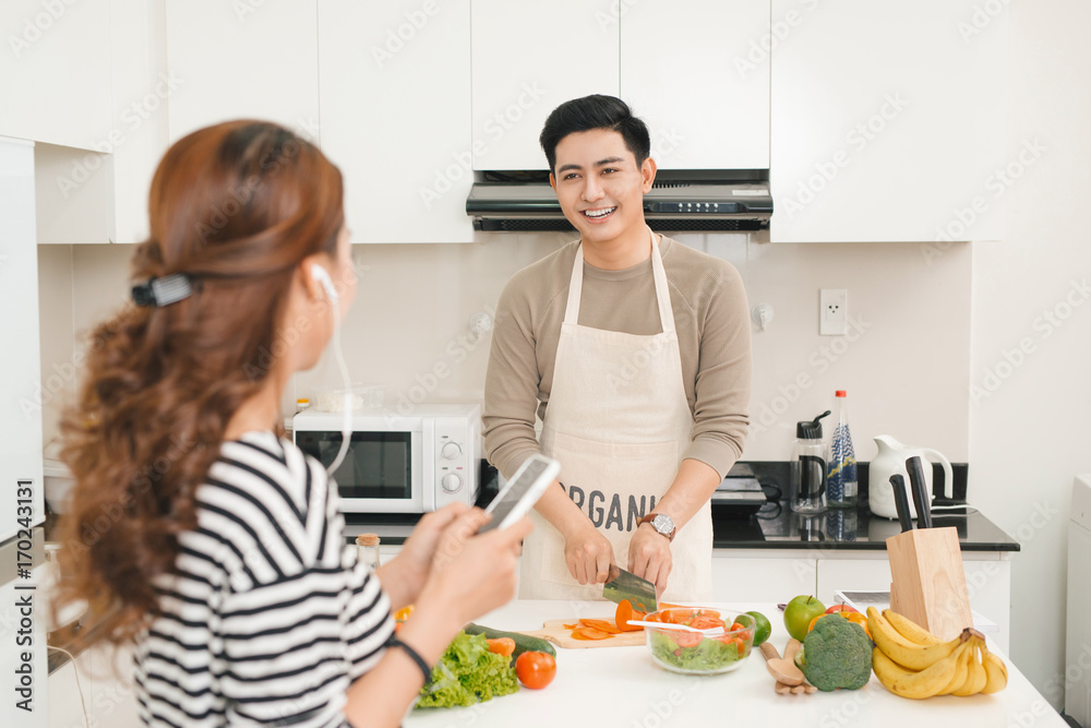 Woman using mobile phone in kitchen as boyfriend prepares meal