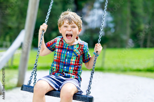 Funny kid boy having fun with chain swing on outdoor playground on sunny day