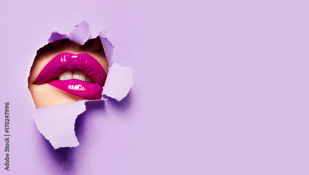 Fototapeta premium Piękne pulchne jasne usta różowego koloru zaglądają w szczelinę kolorowego papieru.