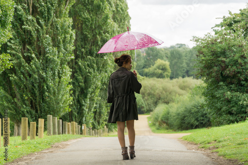 Filipino model under a umbrella walking on a pathway in a park at summertime in the rain, wearing denim shorts a black top with a coat on under a pink umbrella 