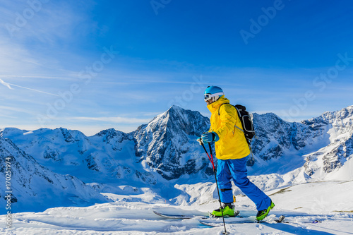 Fototapeta Mountaineer backcountry ski walking up along a snowy ridge with skis in the backpack. In background blue sky and shiny sun and Zebru, Ortler in South Tirol, Italy.  Adventure winter extreme sport.