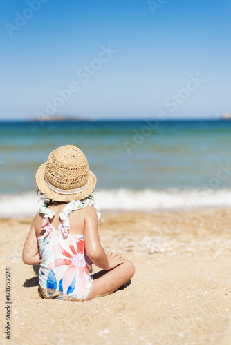 Little girl child with straw hat and floral swimsuit looking at the sea in a sunny day. Little girl sitting back on the shore of the sea