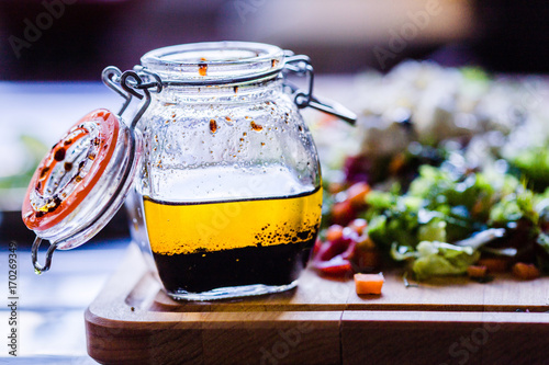 A Glass Bottle with Salad Dressing consisting of Balsamic Vinegar, Olive Oil, Pepper and Salt, Salad on the Background, Horizontal View