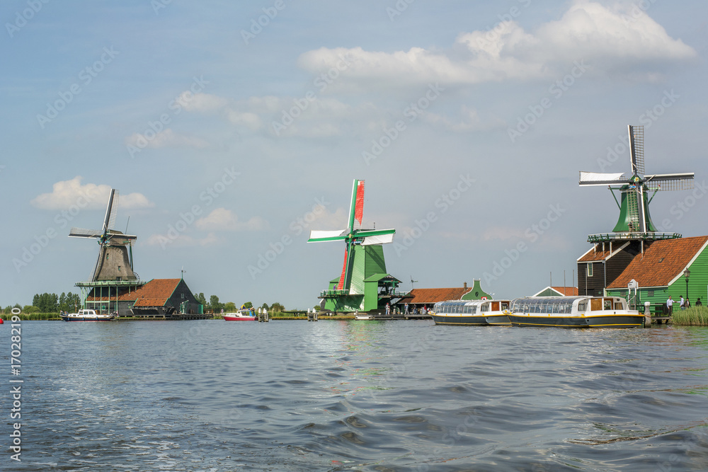 Three mills on the blue water with boats around in the day in Zaanse Schans (Netherlands)