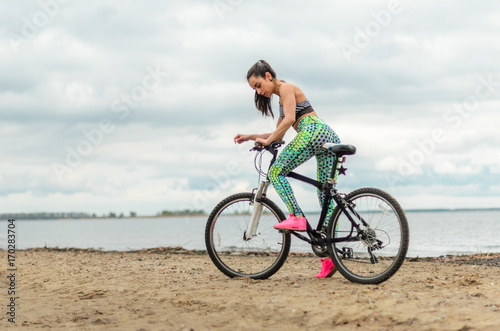Fototapeta Naklejka Na Ścianę i Meble -  The girl the brunette plays sports on the seashore - on the beach. Rides a bike.