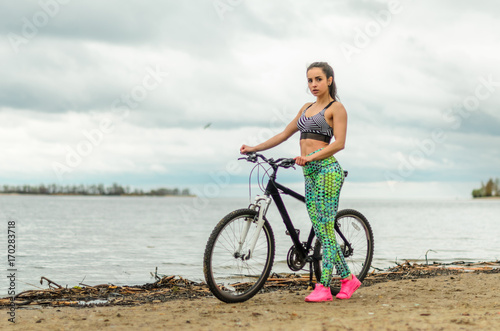 The girl the brunette plays sports on the seashore - on the beach. Rides a bike.