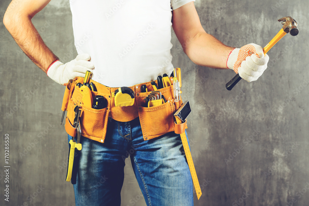unidentified builder standing in white gloves with a tool belt with construction tools and holding a hammer against grey background, toned image. DIY tools and manual work concept. unidentified builder standing in white gloves with a tool belt with construction tools and holding a hammer against grey background, toned image. DIY tools and manual work concept.