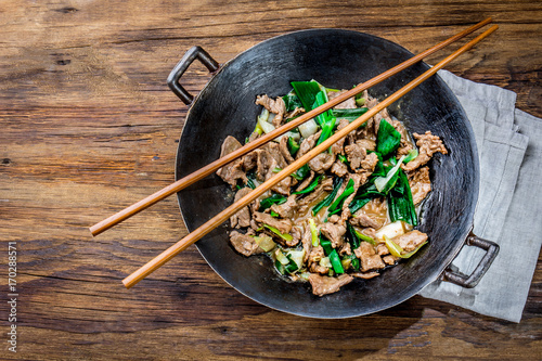 Traditional chinese mongolian beef stir fry in chinese cast iron wok with cooking chopsticks, wooden background. Top view, copy space