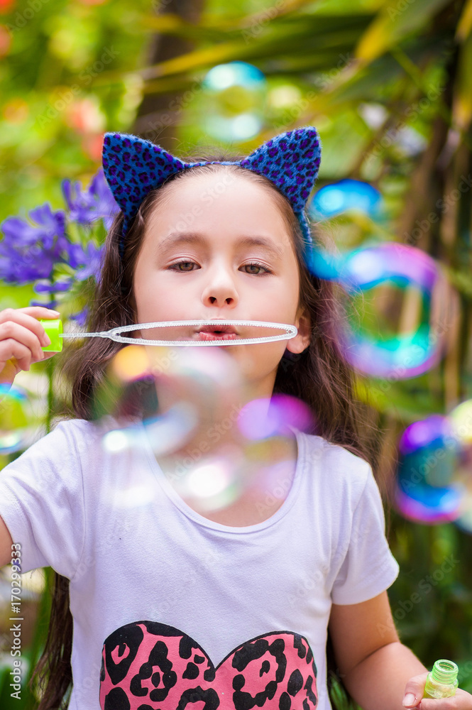 Happy little girl playing with soap bubbles on a summer nature, wearing a blue ears tiger accessories over her head in a blurred nature background Happy little girl playing with soap bubbles on a summer nature, wearing a blue ears tiger accessories over her head in a blurred nature background