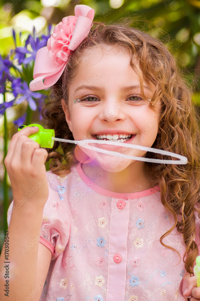 Happy little curly girl playing with soap bubbles on a summer nature, wearing a pink dress and flower in her head, in a blurred nature background Happy little curly girl playing with soap bubbles on a summer nature, wearing a pink dress and flower in her head, in a blurred nature background