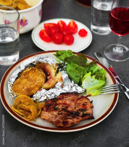 Table with plate of grilled chicken meat, baked potato, salad and tomato, water and vine.