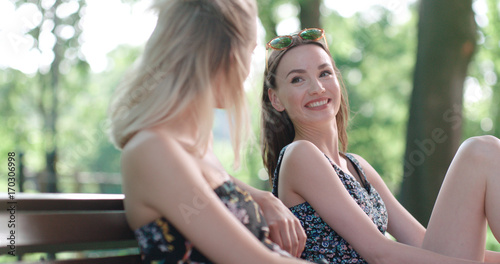 Two young girls sitting on bench in a park enjoying summer and chatting. Positive face expressions, emotions, feelings, body language. 