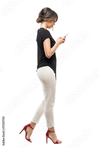 Fototapeta Naklejka Na Ścianę i Meble -  Side view of busy business woman in formal suit walking and typing on mobile phone. Full body length portrait isolated on white studio background