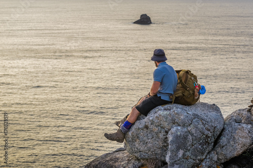 tourist with a backpack sits on a rock looking at the sunset over the ocean