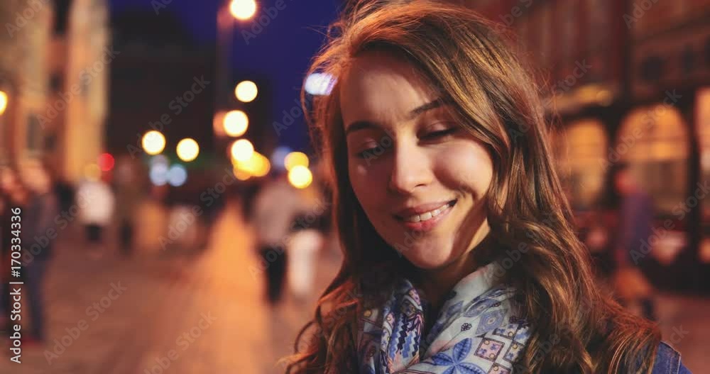 Portrait of a Beautiful Young Woman at Night in the City. SLOW MOTION 4K DCi. Attractive Happy Girl looking to camera. Blurred Night Busy Street lights background. 