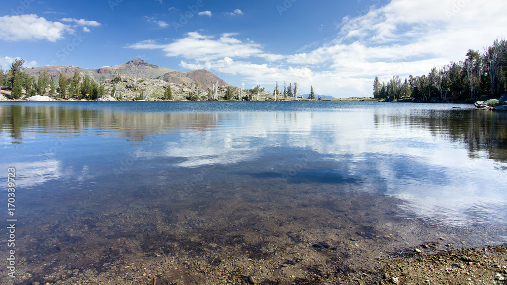 A portion of Sierra Nevada Frog Lake at mid-day in the summer, with rocks,  the mountains and a few pine trees, and clouds in the backgroud
