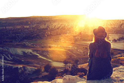 A tourist girl in a hat sits on a mountain and looks at the sunrise.