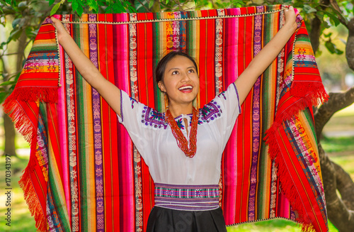 QUITO, ECUADOR - AUGUST, 30 2017: Unidentified young indigenous woman wearing a typical andean clothes, holding with both hands a colorful blanket in the park photo