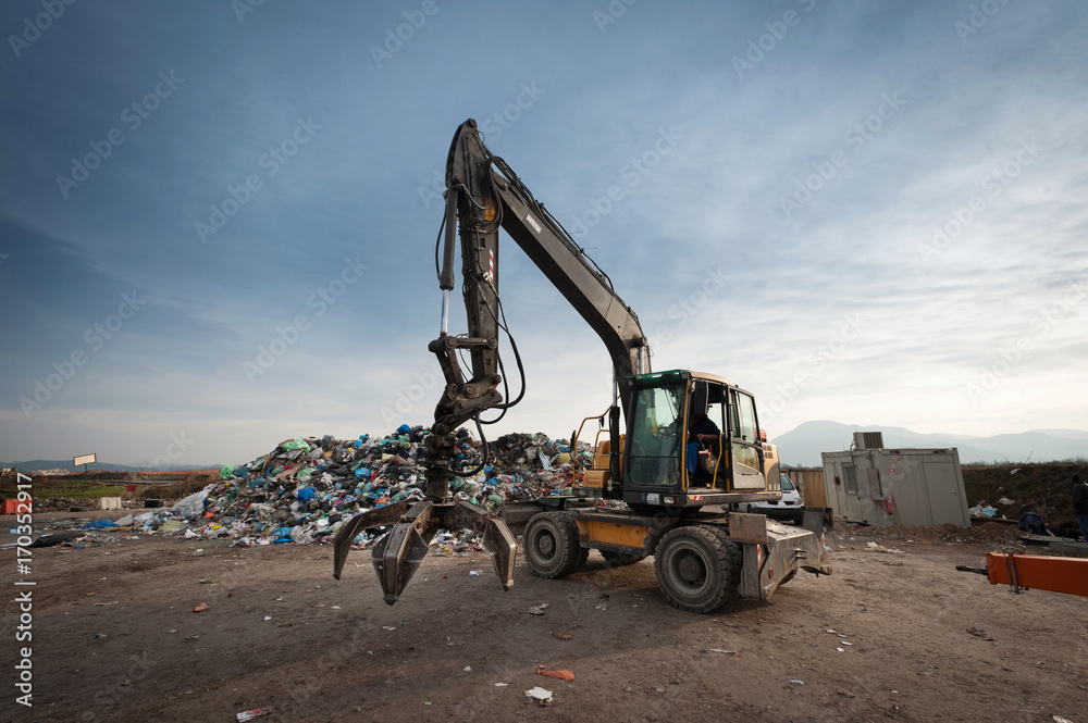 Bulldozer with mechanical arm grabbing waste from a pile at city landfill. Waste management, ecology concept.