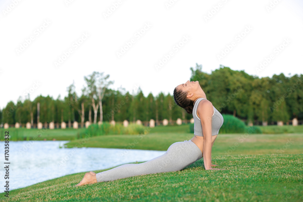 Fototapeta premium Young woman in grey costume practicing yoga near lake