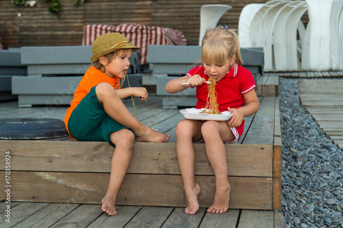 A girl and a boy sitting on a wooden deck and eating noodles